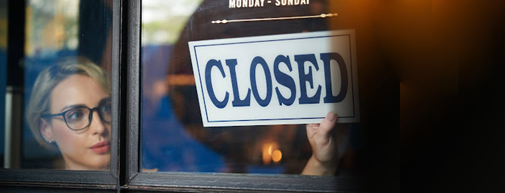 A woman places a 'Closed' sign on the window of her small business, marking a somber moment of company insolvency. This act signifies the difficult decision and impact of financial challenges on her once-thriving establishment.